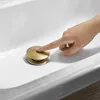 A hand is pressing down on a gold pop-up drain in a white sink. Produktbild-Vorschau