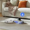 A white robot vacuum cleaner is cleaning a carpeted floor next to a sofa and slippers. A blue cone icon is visible in the upper right corner. Náhled obrázku produktu
