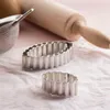 Two oval-shaped fluted tartlet molds are placed on a pink textured surface, with a wooden rolling pin and a sieve in the background. Náhled obrázku produktu
