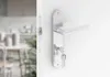 Close-up of a silver door handle with keys inserted, attached to a white door. The background shows a blurred interior of a room with furniture. Produktbild-Vorschau