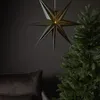 A dark green, seven-pointed star lantern hangs in front of a dark gray wall, with a portion of a green Christmas tree visible on the right and a dark gray cushion on the left. Podgląd zdjęcia produktu