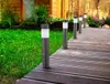 Several solar-powered bollard lights are lined up along a wooden deck next to a grassy lawn and a wooden house. Podgląd zdjęcia produktu