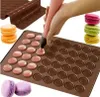 A hand is using a piping bag to fill circular indentations on a brown silicone baking mat, with stacks of colorful macarons visible in the background and foreground. Podgląd zdjęcia produktu