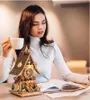 A woman is sitting at a table, holding a cup of coffee and looking at a wooden cuckoo clock model. Náhľad obrázku produktu
