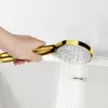 A hand holding a gold shower head over a bathtub, with water spraying from the shower head. Produktbild-Vorschau