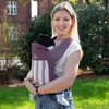 A woman is holding a baby in a carrier outdoors. Produktbild-Vorschau