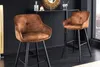 Two brown bar stools with black metal legs are placed in a modern, dimly lit room with a black wall and dark flooring. Produktbild-Vorschau
