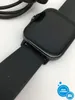 Close-up of a black smartwatch with a textured strap, with charging cables in the background. Voir les images du produit