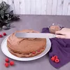 A hand is using a spatula to spread chocolate frosting on a cake, with raspberries scattered around. Produktbild-Vorschau