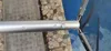 A close-up view of a silver pole with two buttons, attached to a blue net with a gray frame. The background shows a stone-tiled area and a blue surface, possibly a pool. Produktbild-Vorschau