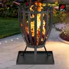 A black metal fire pit with flames burning inside, set on a paved patio with grass and plants in the background. Produktbild-Vorschau