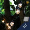 String lights with clear bulbs hanging from tree branches, with a solar panel in the lower right corner. Produktbild-Vorschau