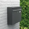 A dark gray mailbox mounted on a white brick wall with a blurred green background of foliage. Produktbild-Vorschau
