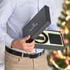 A person holding a gift box containing a belt and wallet, with a Christmas tree in the background. Anteprima dell’immagine prodotto