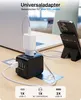 A universal travel adapter is shown plugged into a laptop, with a phone charging on a stand behind it. The adapter has multiple USB ports and a universal AC socket. Náhled obrázku produktu