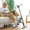 An elderly man sits on a couch reading a book while using a pedal exerciser for his legs. Produktbild-Vorschau