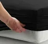 A hand is shown tucking a black fitted sheet onto a mattress. Produktbild-Vorschau
