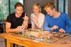 Three people are playing a board game at a wooden table. Náhľad obrázku produktu