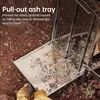 A close-up of a camping stove's pull-out ash tray filled with ash, placed on the ground. Náhled obrázku produktu