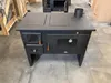 A black wood-burning stove with a glass oven door and stovetop is shown in a warehouse or workshop setting. Náhľad obrázku produktu