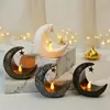 Several moon-shaped candle holders with LED lights and small stars are displayed on a marble surface, with blurred fairy lights in the background. Produktbild-Vorschau