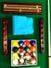 A collection of billiard balls, cue rack, and scoring board arranged on a green billiard table. Produktbild-Vorschau