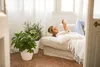 A woman is relaxing on a couch, reading a book, surrounded by plants. Náhled obrázku produktu