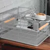 A gray metal dish drying rack with a wooden handle utensil holder is shown on a wooden surface against a white brick wall. Náhľad obrázku produktu