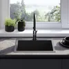A modern kitchen sink with a black faucet, set against a window with a view of greenery and white flowers. Produktbild-Vorschau