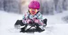 A young child is lying on a sled in the snow, wearing a pink helmet and goggles. Anteprima dell’immagine prodotto