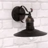 A wall-mounted outdoor light fixture with a dark metal finish and a clear glass shade, mounted on a white brick wall. Anteprima dell’immagine prodotto