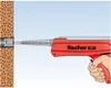 A hand is using a red Fischer injection mortar gun to insert a metal anchor sleeve into a brown, textured wall. Voir les images du produit