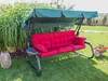 A garden swing with red cushions is set up on a green lawn, surrounded by lush greenery and a few potted flowers. Náhled obrázku produktu