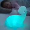A child sleeping in bed with a glowing dinosaur night light on a table in the foreground. Voir les images du produit