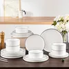 A set of white ceramic dishes with black trim is arranged on a wooden table, with a vase of flowers and a coffee maker in the background. Produktbild-Vorschau