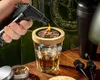 A hand is using a torch to light wood chips in a glass for smoking a drink. Voir les images du produit