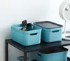 Two turquoise storage boxes with black lids are placed on a black shelf. One box contains a remote control and a pair of headphones. A white fan is visible in the background. Produktbild-Vorschau