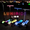 Four LUMI RIDE scooters with LED wheels are lined up in the dark, with city lights in the background. Produktbild-Vorschau