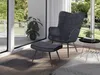 A dark gray velvet armchair and ottoman are placed next to a large window with a view of greenery. A small black side table with a decorative object sits to the right of the armchair. Produktbild-Vorschau
