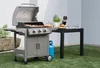 A gas grill is set up on a lawn next to a table with food and utensils. Náhled obrázku produktu