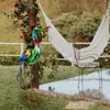 A decorative metal peacock statue stands in a garden next to a wooden post with vines and a white hammock in the background. Produktbild-Vorschau