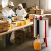 Two beekeepers in protective suits are processing honey. One is holding a honeycomb, and the other is operating a honey extractor. Jars of honey are lined up on a table. Produktbild-Vorschau