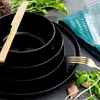 A stack of black plates and bowls with gold cutlery, set on a dark surface with fresh basil and garlic. Náhled obrázku produktu