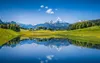 A serene mountain landscape with a lake reflecting the snow-capped peaks and green hills under a clear blue sky. Náhled obrázku produktu