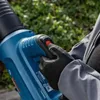 A person wearing gloves is holding a blue leaf blower outdoors. Produktbild-Vorschau