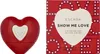 A red heart-shaped perfume bottle and its packaging are displayed side-by-side against a white background. Podgląd zdjęcia produktu