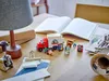 A collection of LEGO® building blocks, including a moving truck, figures, and furniture, arranged on a wooden table with an open book and a lamp. Náhľad obrázku produktu