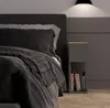 A close-up view of a modern bedroom with a dark gray upholstered headboard, dark gray bedding, and a small gray nightstand with books. Náhled obrázku produktu