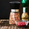 A close-up shot of a glass salt grinder filled with pink salt and spices, with a small glass bowl of red peppercorns in the foreground and another spice grinder in the background. Produktbild-Vorschau