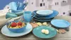 A collection of blue and teal ceramic dishes, including bowls, plates, and cups, arranged on a wooden surface with seashells. The background shows a blurred image of white buildings with blue accents, suggesting a Mediterranean or coastal theme. Produktbild-Vorschau